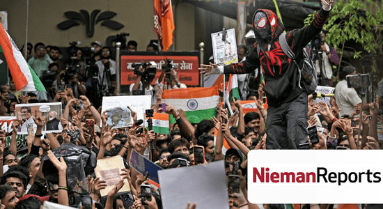 A large crowd. A person with a Spiderman hoodie and mask stands over the crowd with their arms out. People hold posters and the Indian national flag. Others hold up phones on selfie sticks. A press crew with large cameras records the large gathering from near a building. Bottom right, the logo for Nieman Reports. 